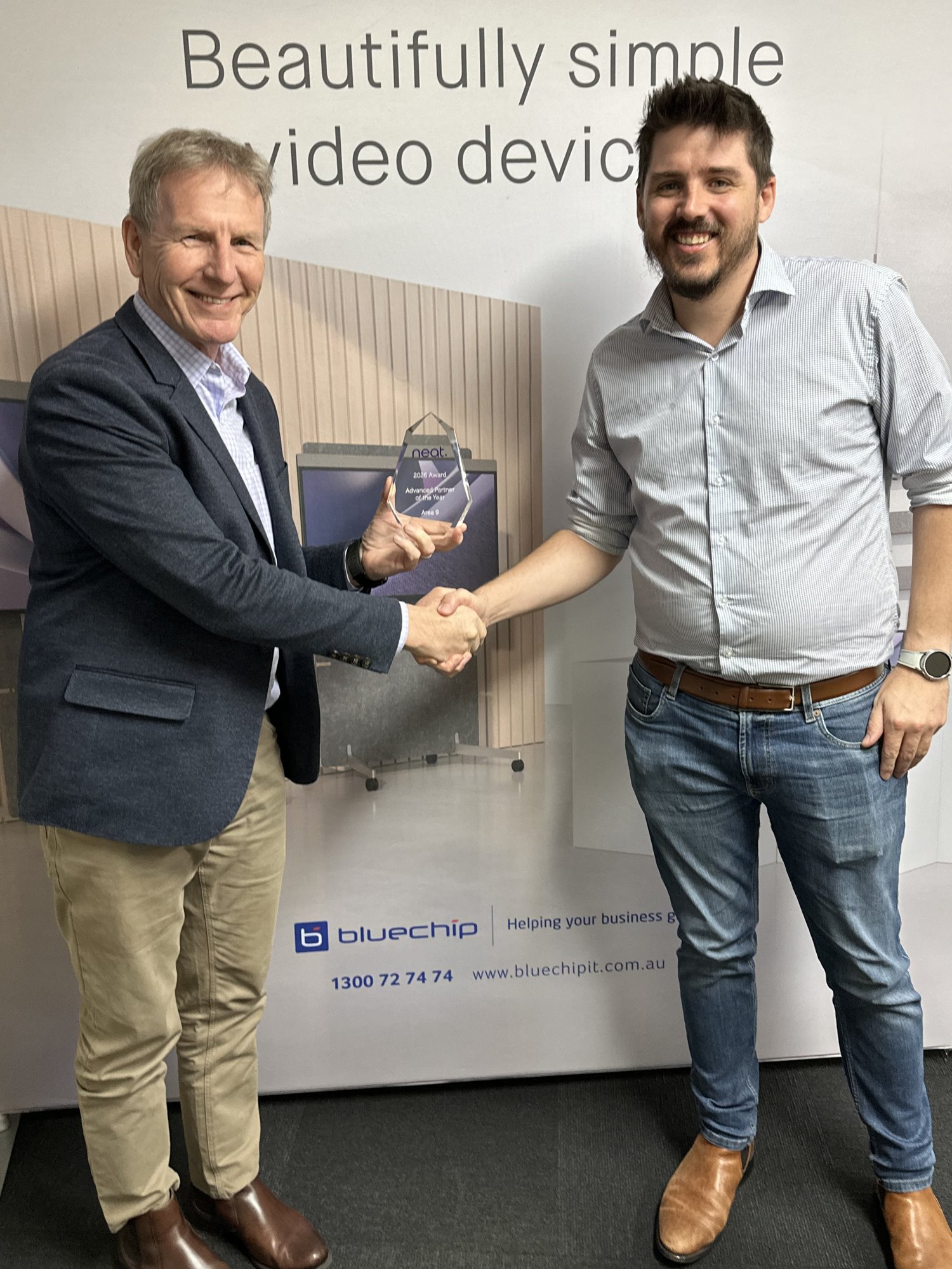 Simon Watt of Area9 holds a glass Neat award and shakes hands with Shane Edwards of Bluechip Infotech in front of a Neat display
